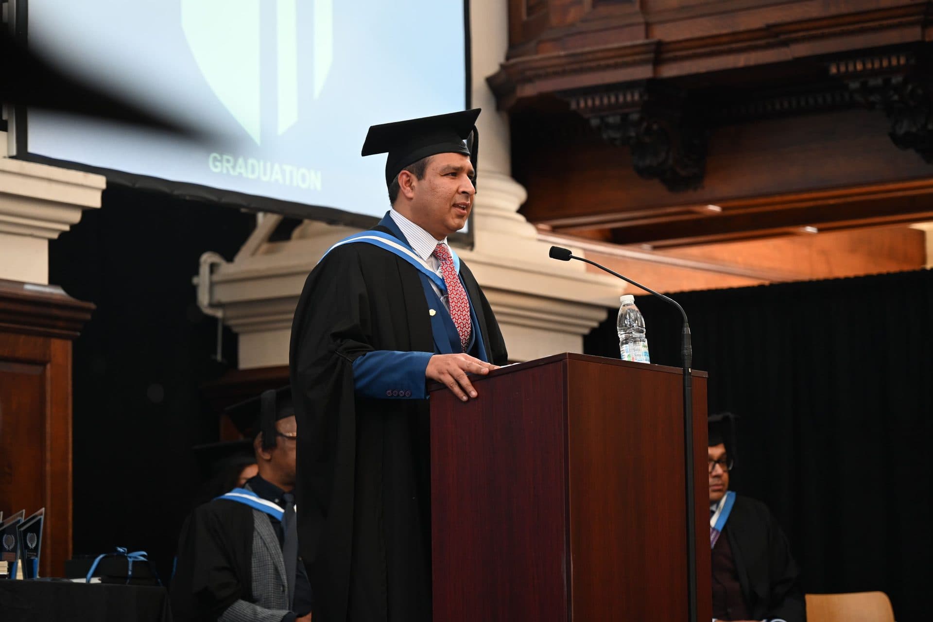 A speaker in academic gown and hood addressing the Class of 2024 ceremony from a lectern