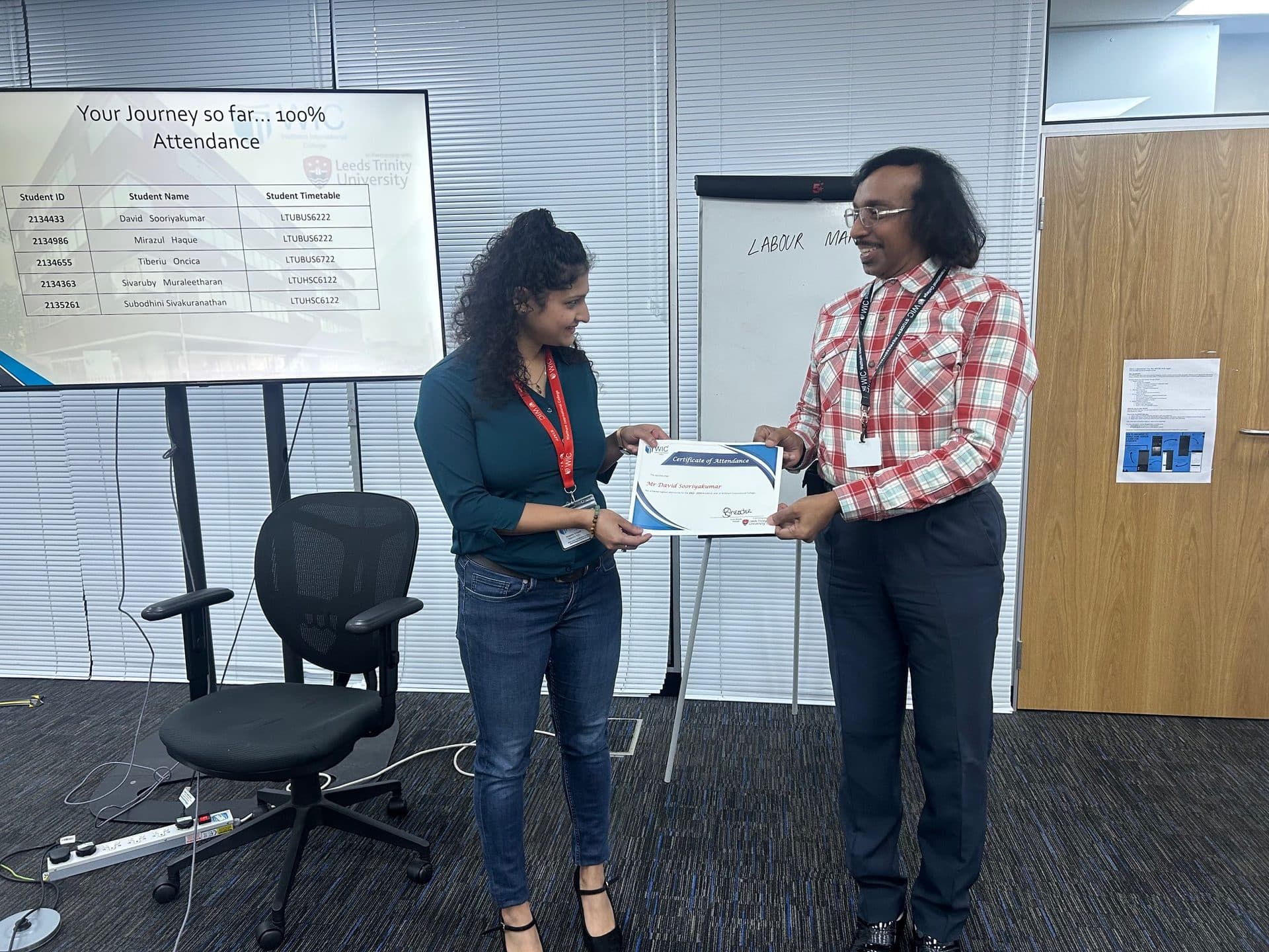 A WIC staff member presenting a Certificate of Attendance to a student, with a '100% Attendance' screen in the background