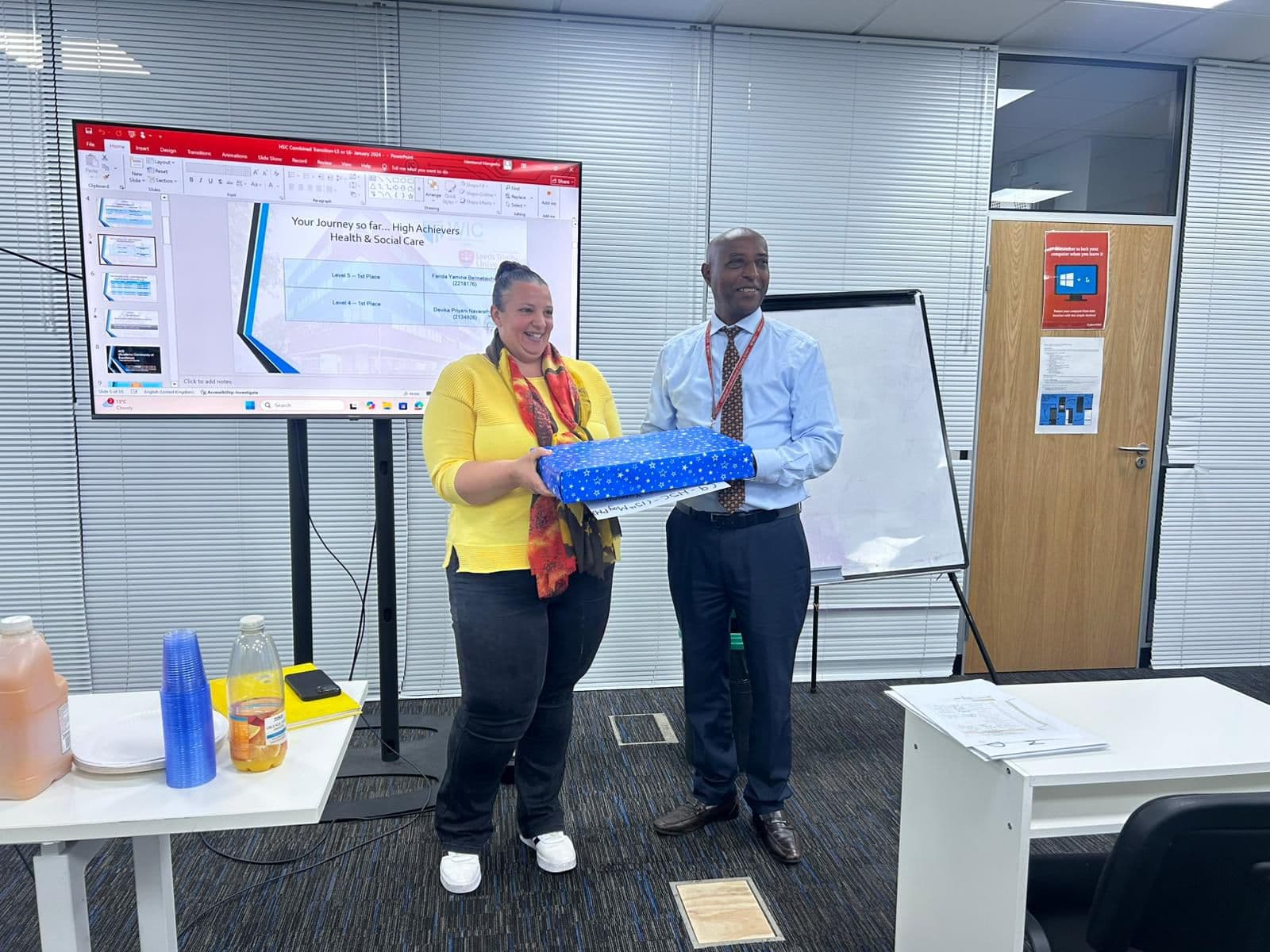 A WIC staff member presenting a wrapped award to a student, with a High Achievers presentation on screen